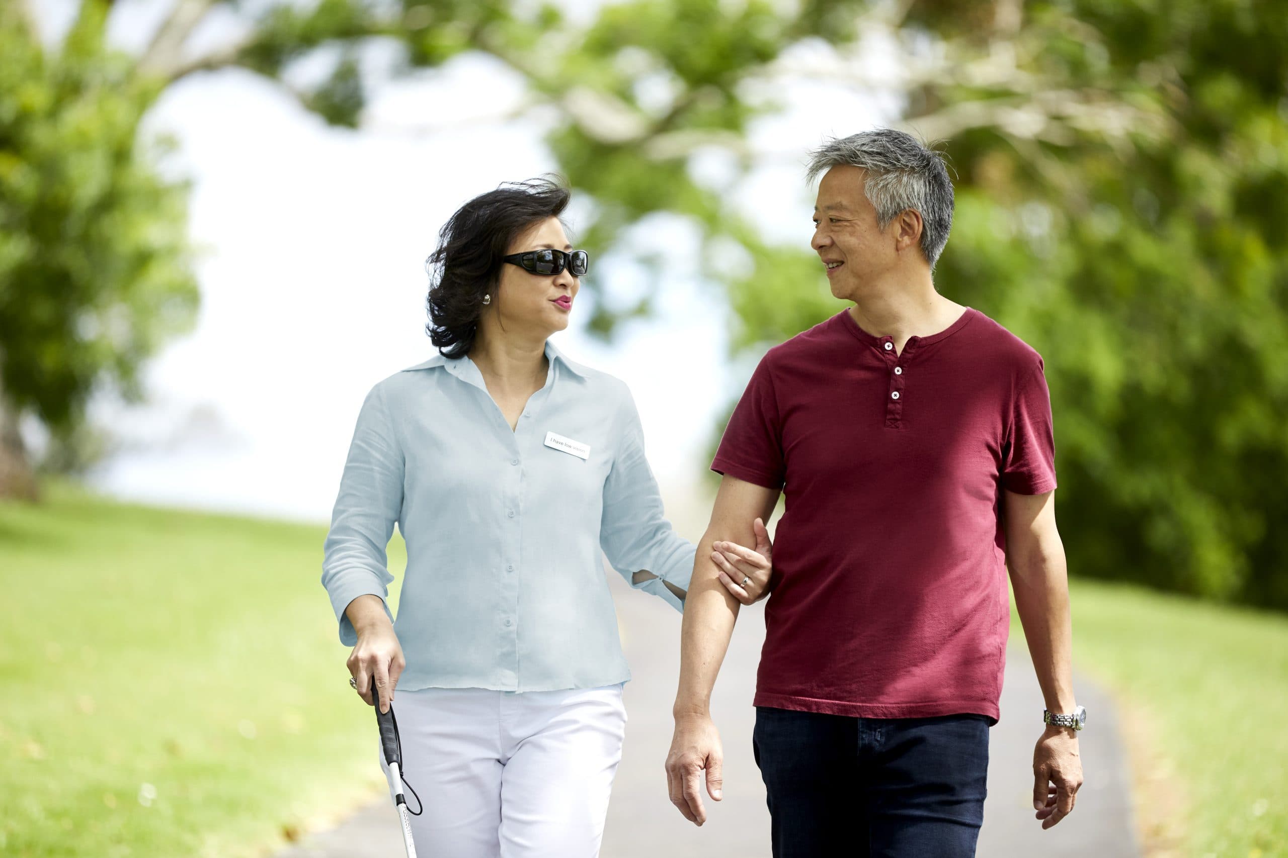 Client Karen and her partner are walking through the Auckland Domain on a bright day, she holds a white cane in one hand and his arm in her other.