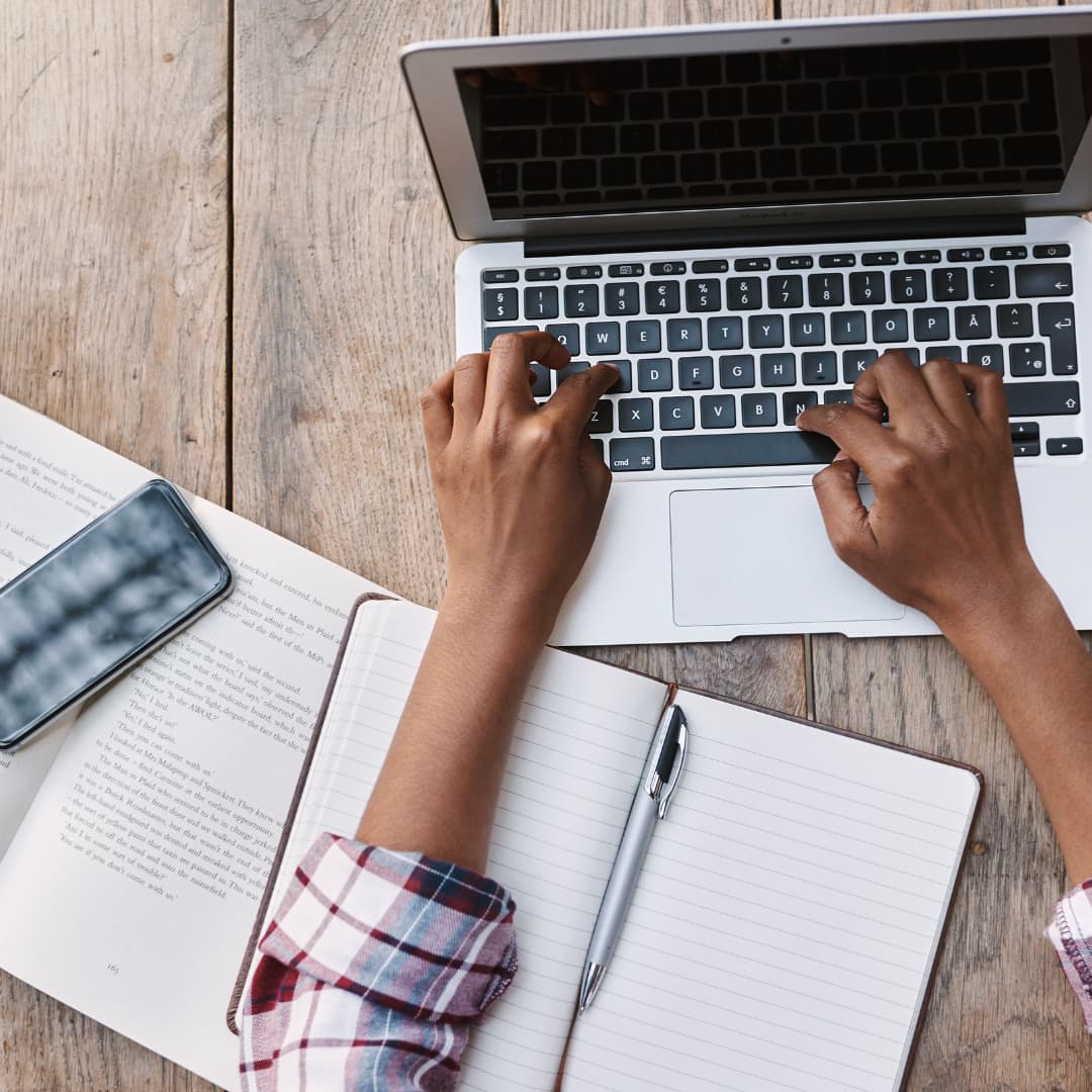 An image of a desktop with a laptop computer, some paper notebooks and a pen. There is a pair of typing on the laptop keyboard.