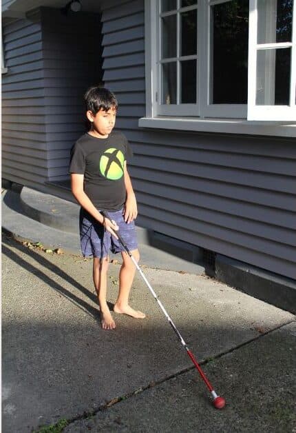 A young blind boy walking outside his house with a white cane.