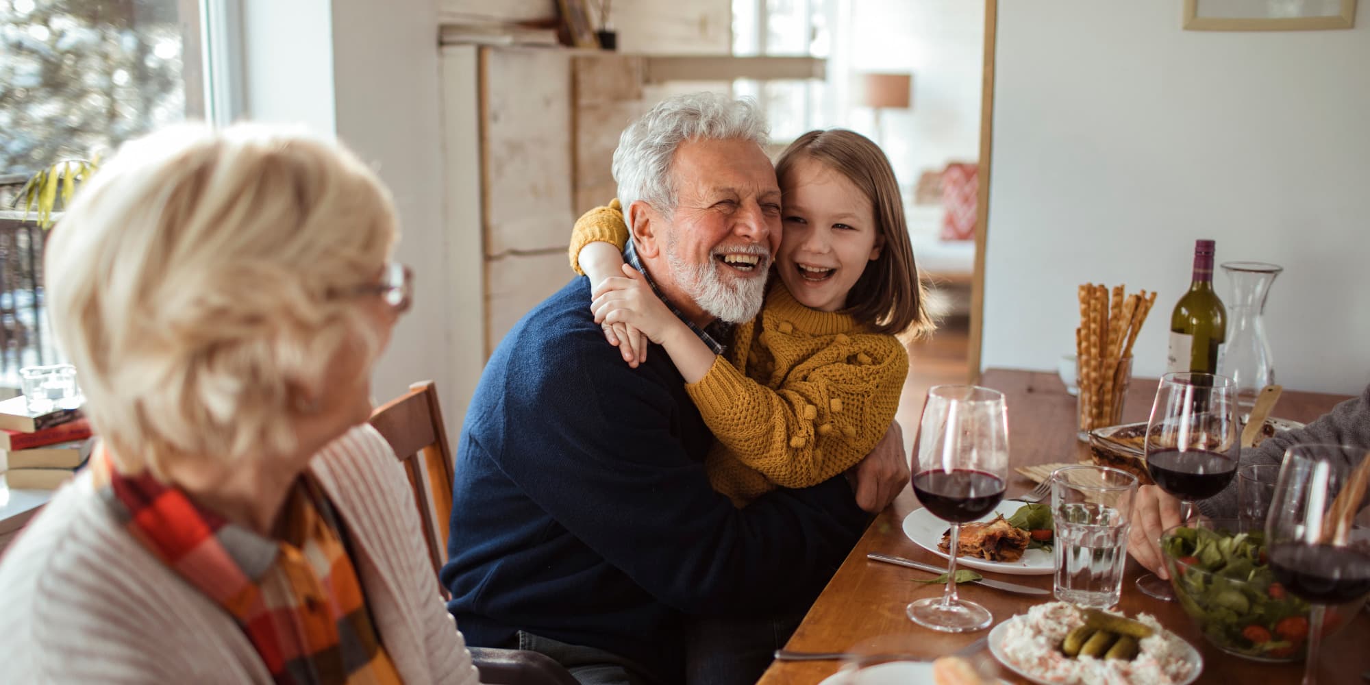 A grandfather laughing as a granddaughter hugs him at the dinner table with the grandmother looking on with hippieness.