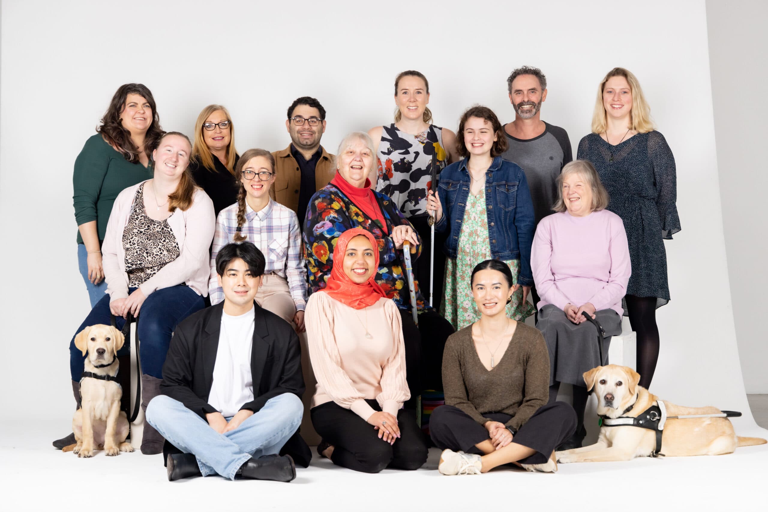 A diverse group of 14 people and two guide dogs sitting at either end of the group.