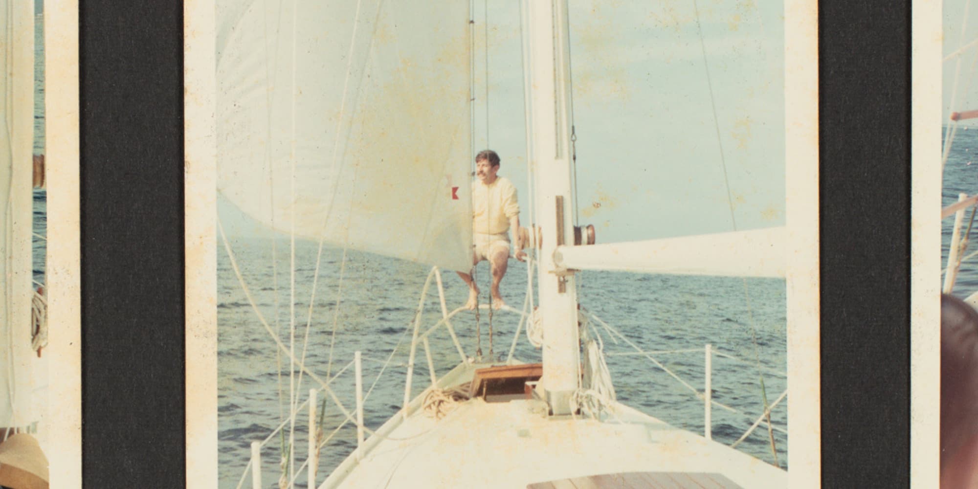 A person stands on the deck of a sailing boat, holding onto a rope, with the ocean and a clear sky in the background. The boat has a wooden deck with various riggings, and the image has the appearance of an old photograph with wear marks around the edges.