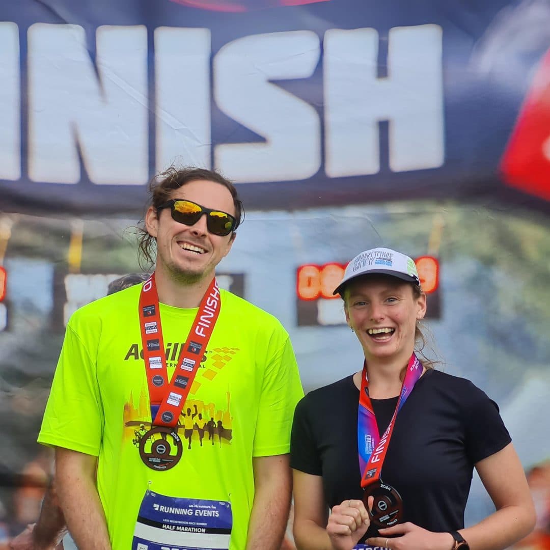 James, a blind runner, standing at the finish line of an event with featured young volunteer, Lydia White. They're both wearing finisher medals and smiling for the camera.