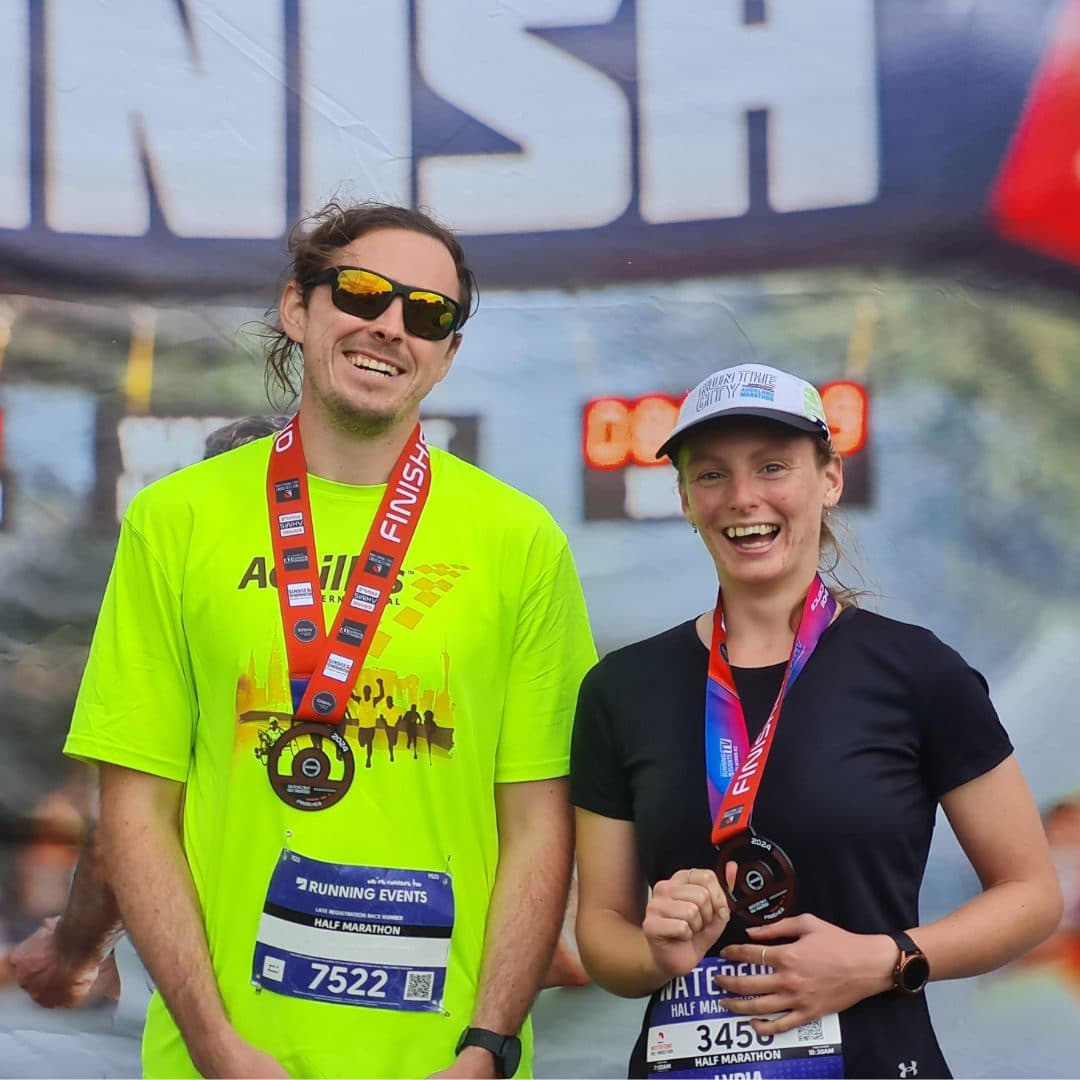James, a blind runner, standing at the finish line of an event with featured young volunteer, Lydia White. They're both wearing finisher medals and smiling for the camera.