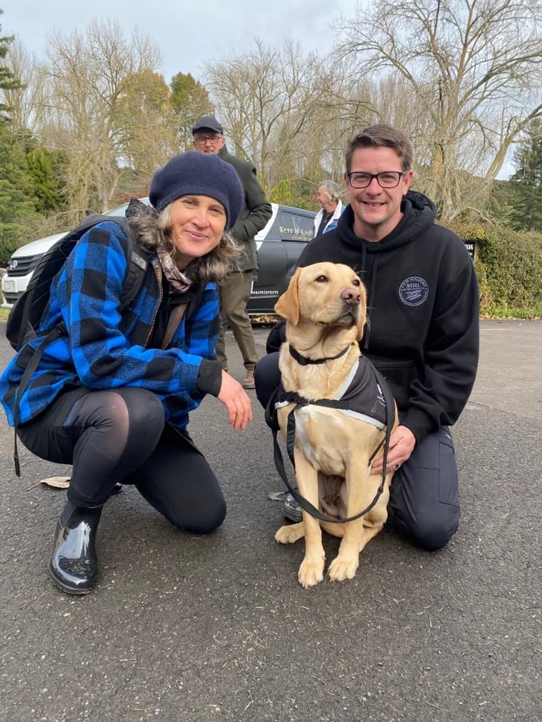 A woman with blonde hair and a wool hat, and a man with short brownm hair and glasses squat next to a Blind Low Vision NZ Guide Dog in a branded harness outdoors in a carpark. Both man and woman are smiling for the camera.