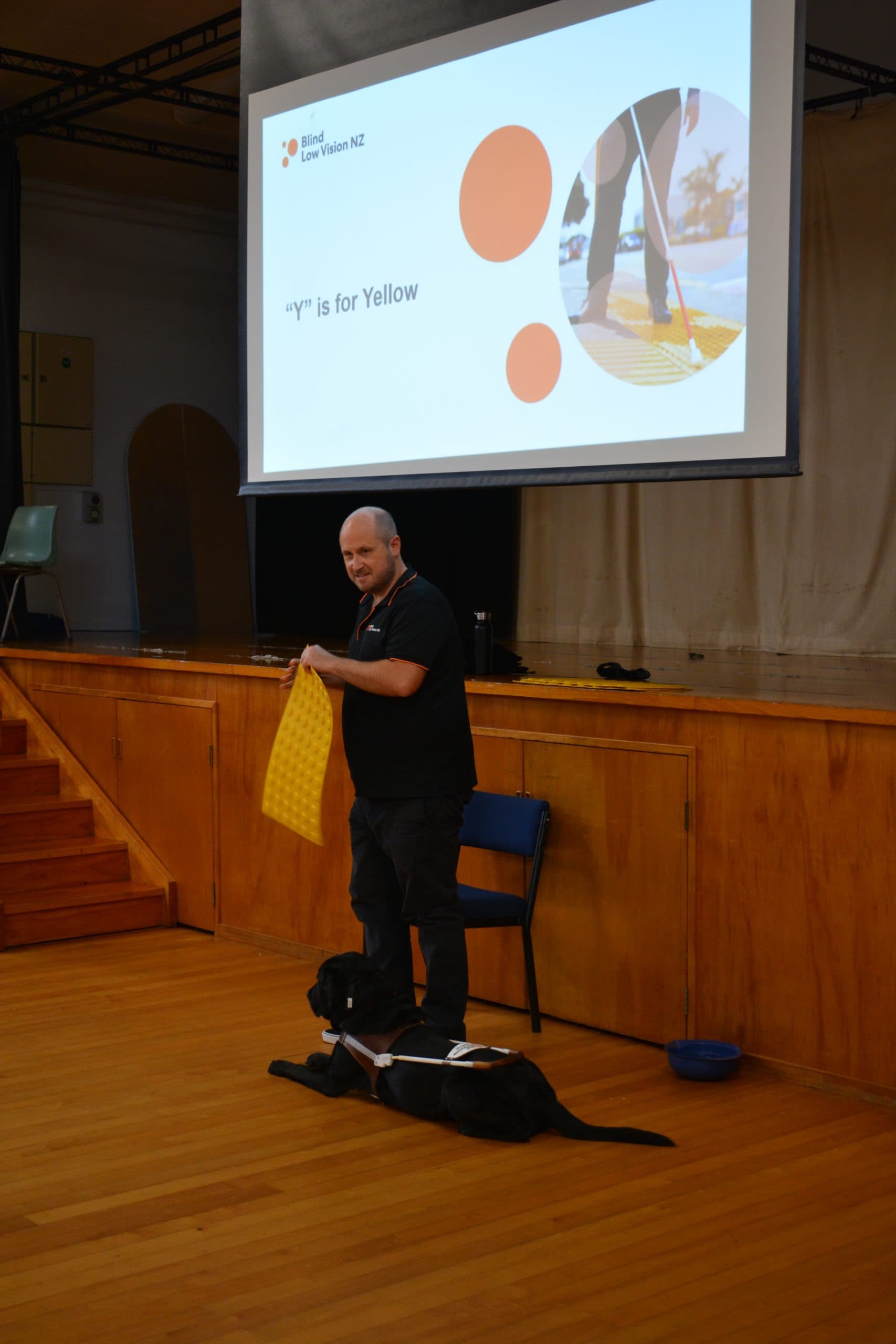 Dan Shepherd, Head of Community Engagement at Blind Low Vision NZ in a black Blind Low Vision polo and black pants standing in a school hall with his guide dog Ezra at his feet. Dan is holding some yellow tactile dots that can be found on a lot of pedestrian walkways.