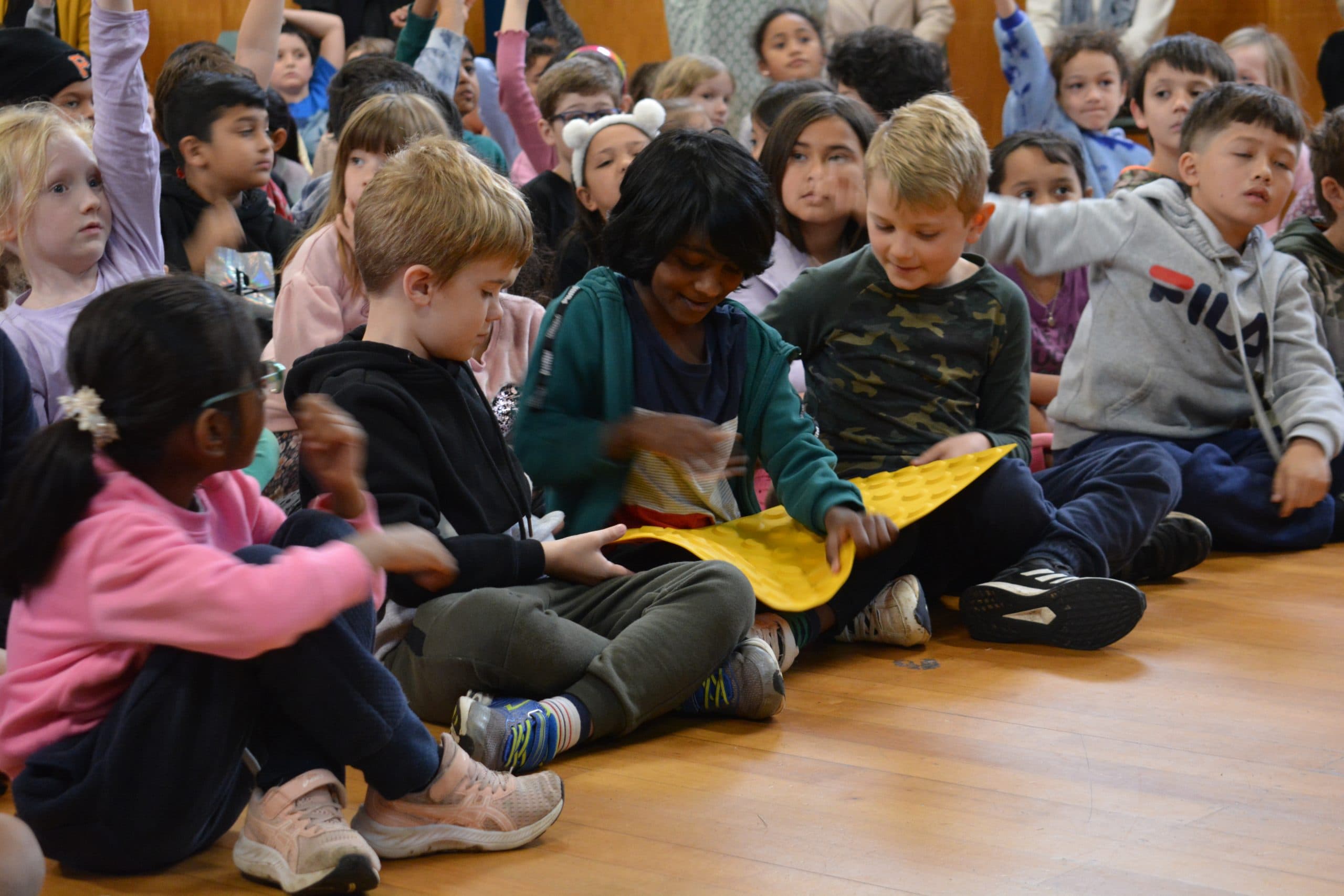 A group of children gathered around a young boy who is seeing what the yellow tactile pedestrian crossing dots feel like.