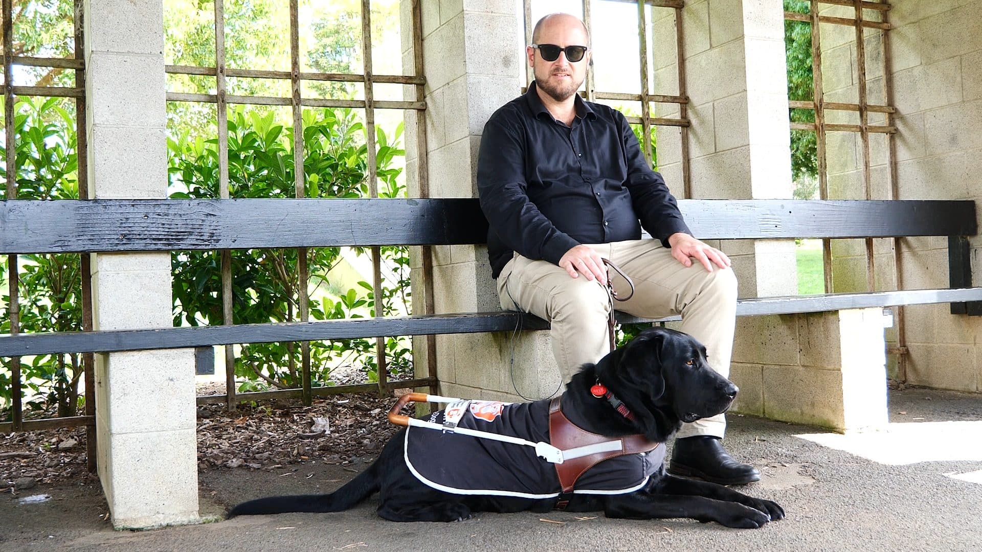 A man wearing dark glasses sitting on an outdoor bench with a black labrador retriever at his feet, wearing a jacket with Blind Low Vision NZ Guide Dogs branding.