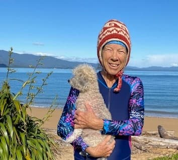 A woman holding her dog at a beach with a flax bush to the left and mountains in the distance. It's a clear blue day.