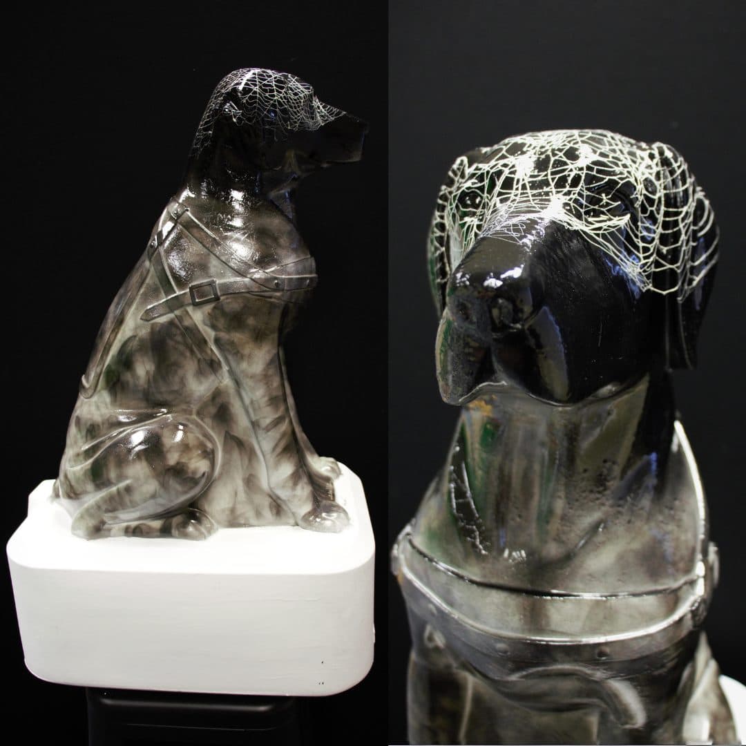 A repurposed Blind Low Vision NZ Trudy dog with a glossy, metallic finish, displaying intricate textures and patterns over its body, positioned on a white pedestal against a black background.