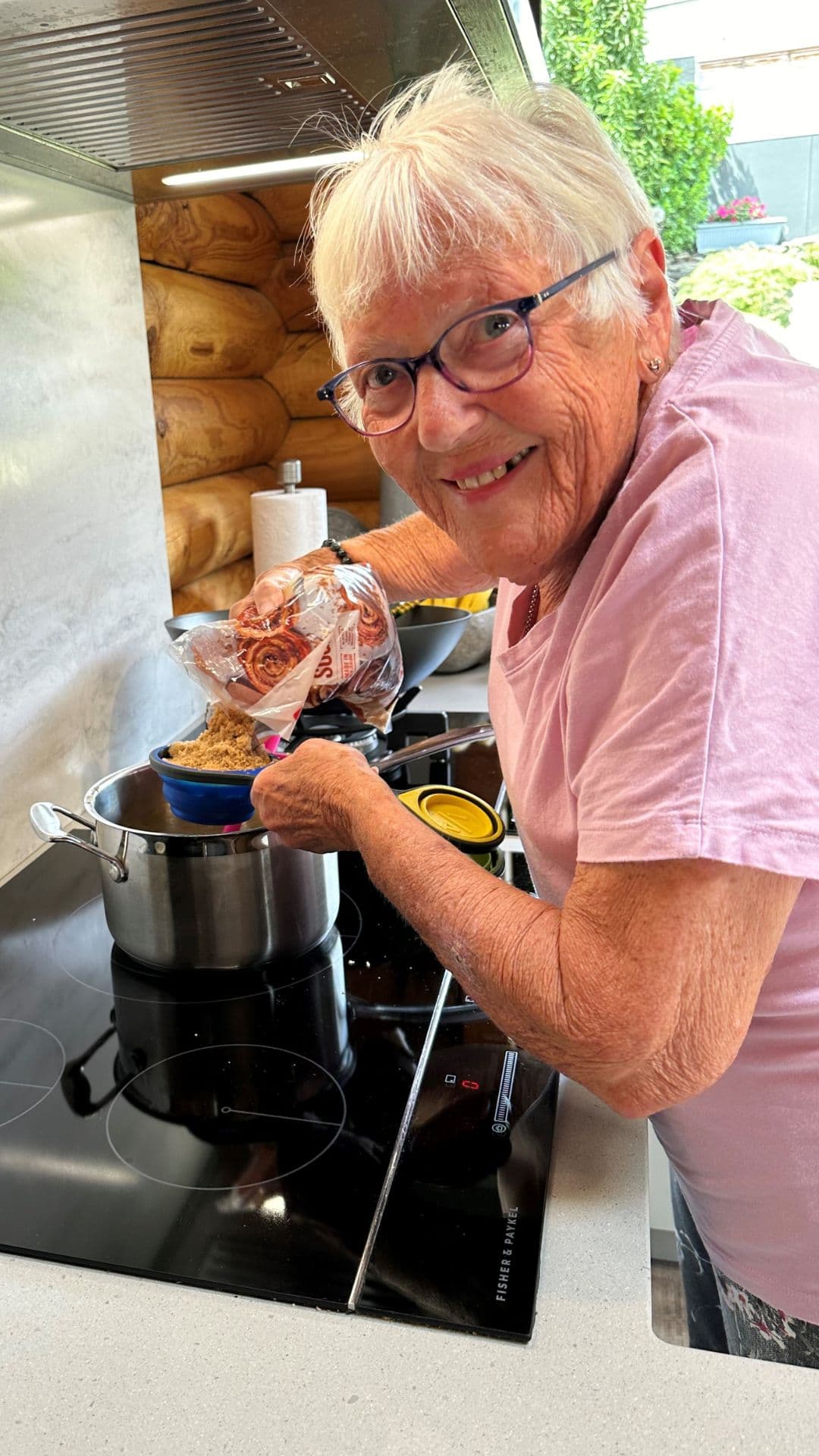 Ellice is standing at her stove top, pouring brown sugar into a measuring cup above a pot on the stove. She is smiling, wearing glasses and a soft pink t-shirt.