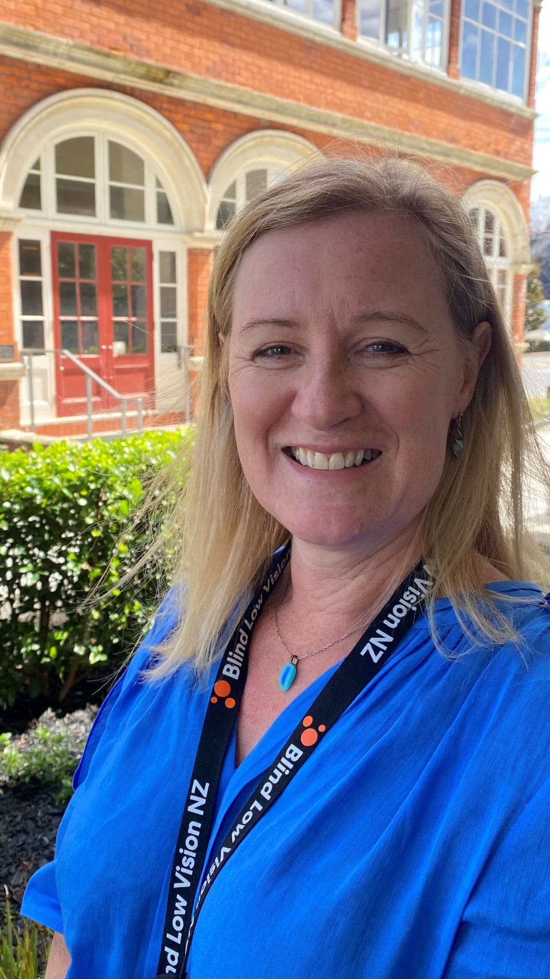 Joanne is taking a selfie, smiling in front of a short hedge with a building behind. She has shoulder length blonde hair and is wearing a vibrant blue top, a blue pendant necklace and a Blind Low Vision NZ lanyard.