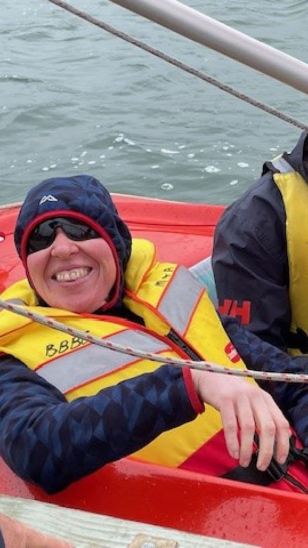 Neroli is smiling, sitting back in a small sailboat, wearing a dark blue athletic jacket with the hood up, a bright yellow life jacket and black sunglasses. Sitting next to her in the boat there is a smiling man wearing a black raincoat underneath a yellow life jacket.