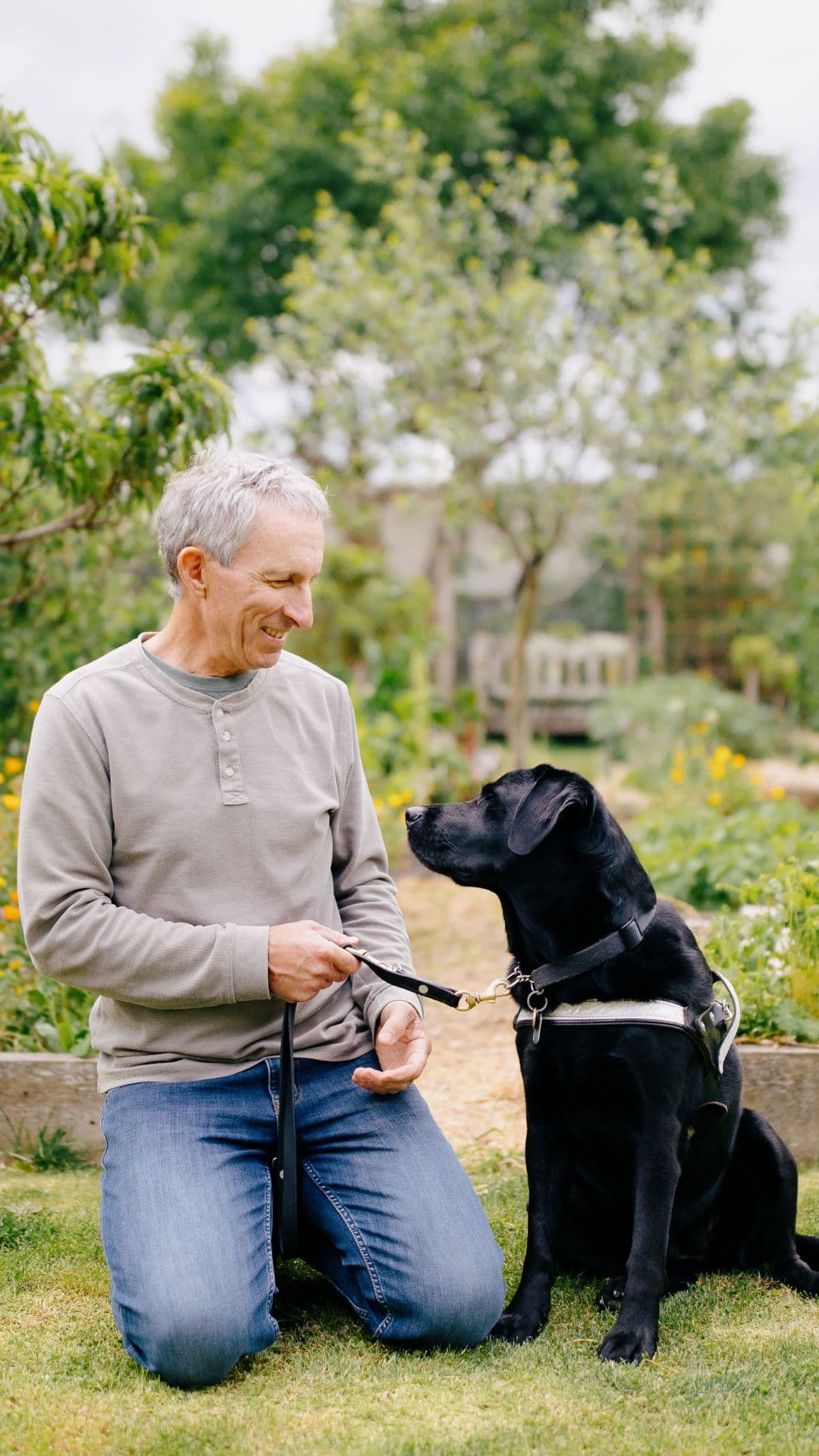 Peter is kneeling down in a garden amongst some flower beds, with his black labrador guide dog Ayla sitting next to him. Peter is smiling and holding Ayla’s leash, wearing a grey long sleeve top and blue jeans.