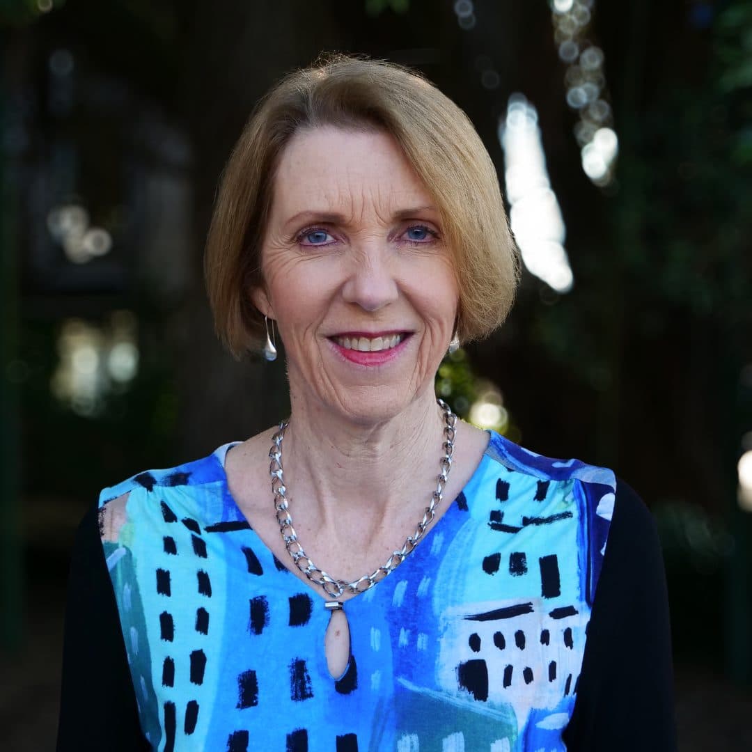 Andrea Midgen, Chief Executive of Blind Low Vision NZ outdoors, smiling at the camera with a tree in the background. Andrea is wearing a blue, white and black patterned top.