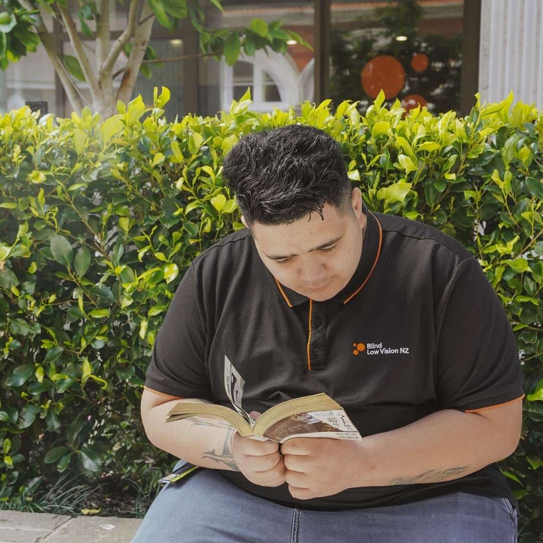 A man in a black t-shirt sitting down outside reading.