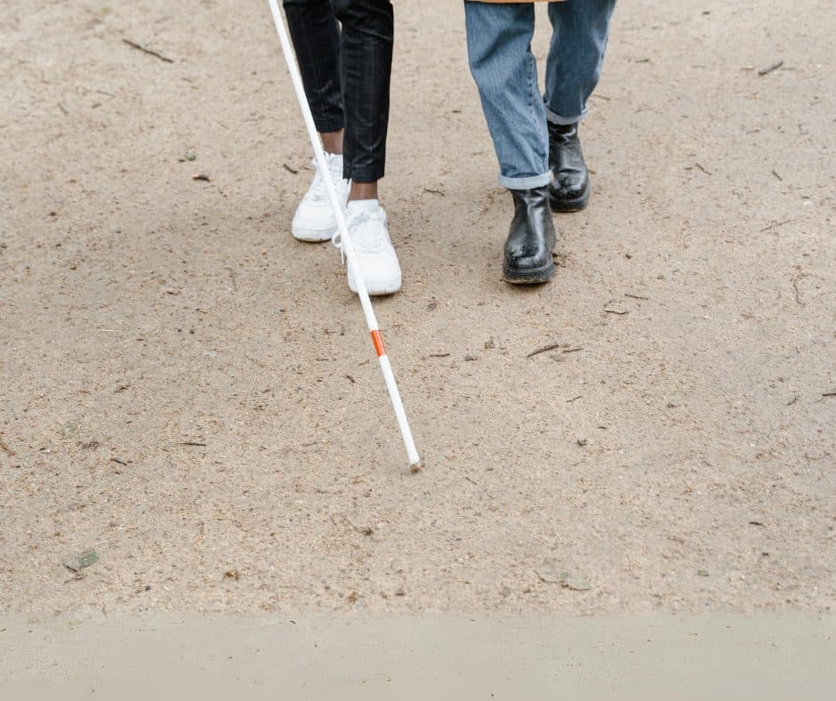 Two people walking, one with a white cane on a beach. Photo shot from the knees down.