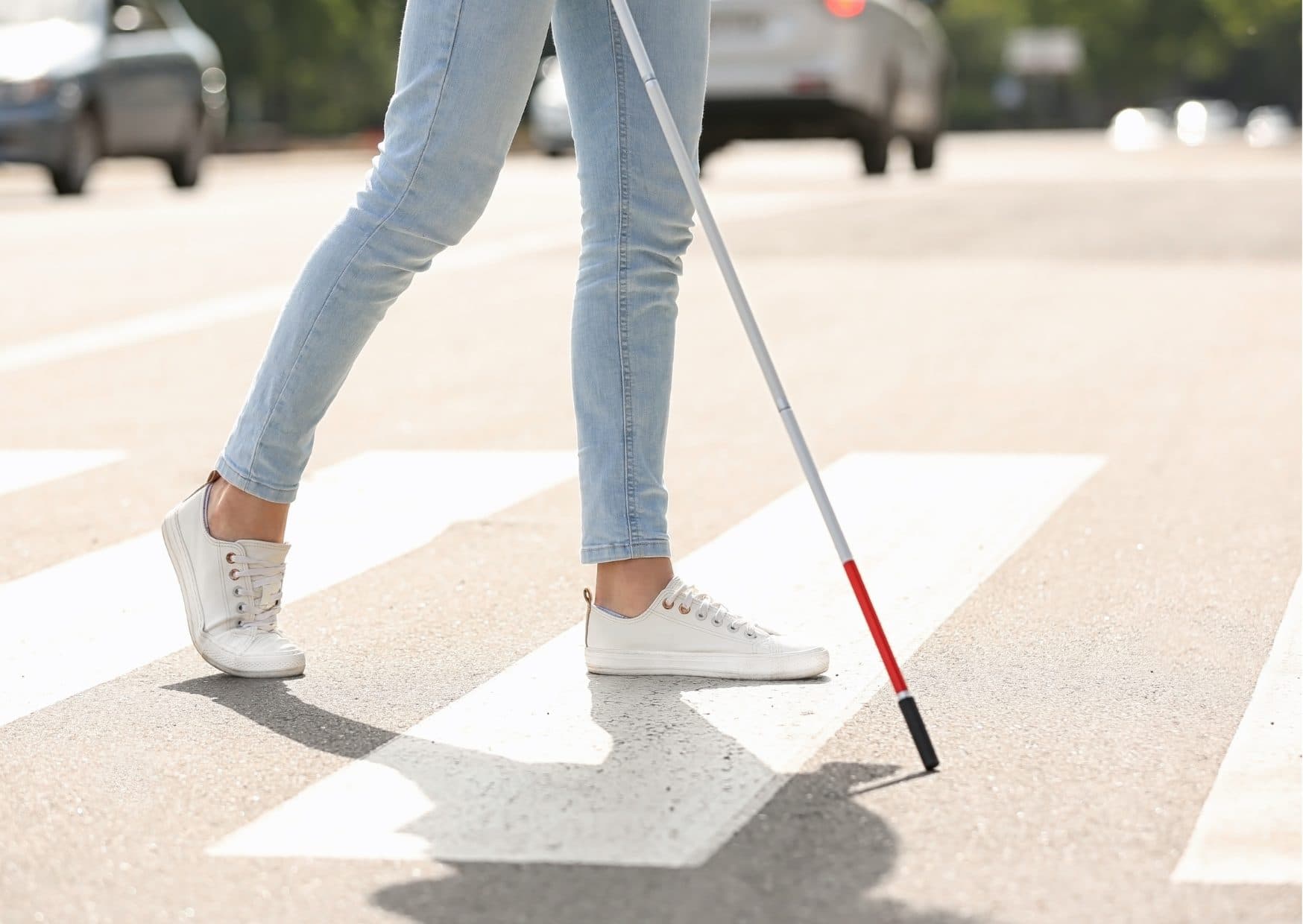 A street level view of a woman in blue jeans and white sneakers on a pedestrian crossing with a white cane.