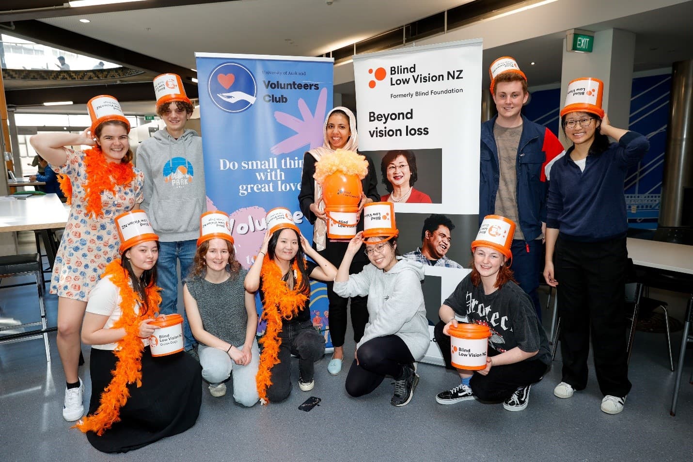 A group of Auckland University Student volunteers gathered around a Blind Low Vision NZ promotional display, posing with orange collection buckets.