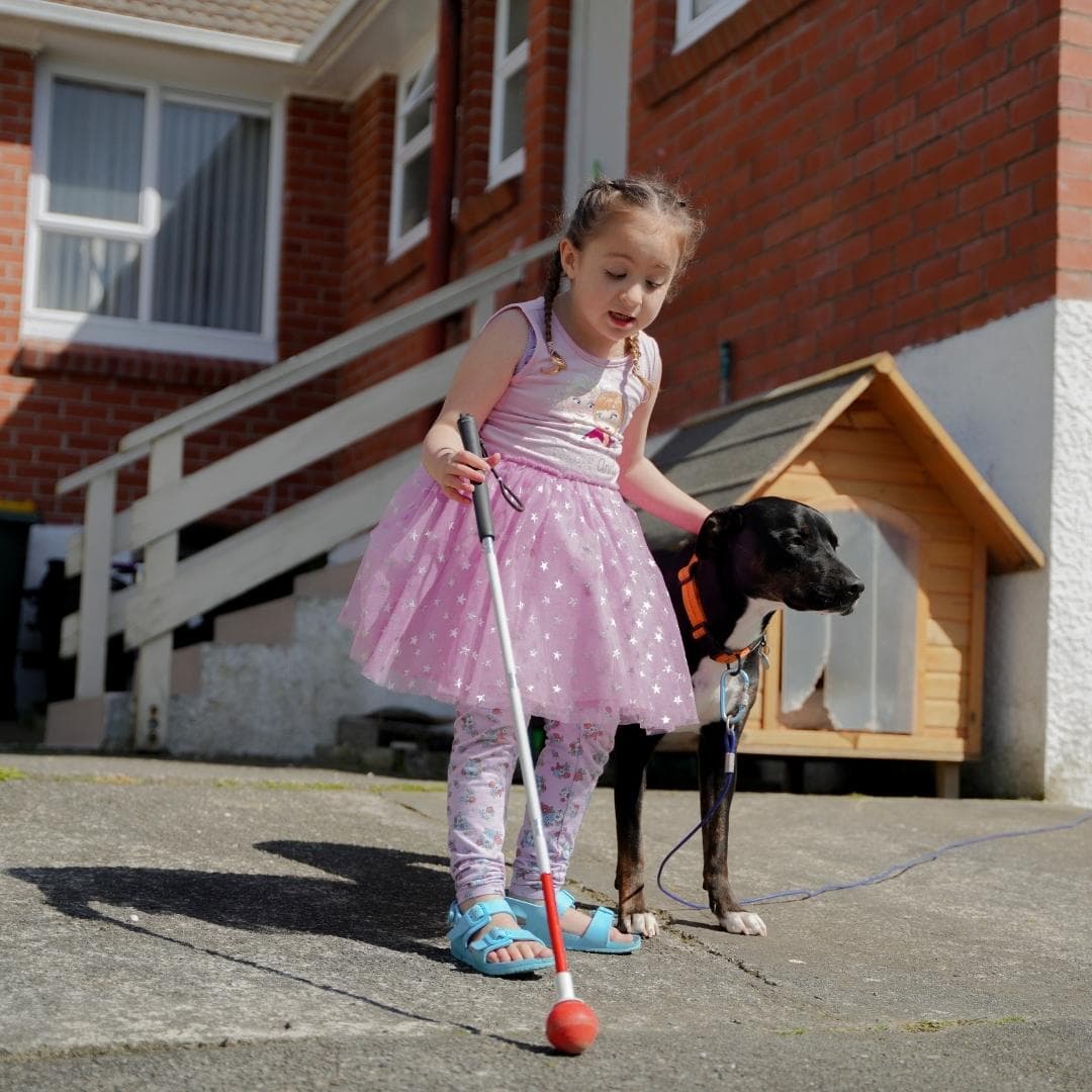 A child dressed in a tutu with her white cane and dog. She is standing on the driveway outside her house.