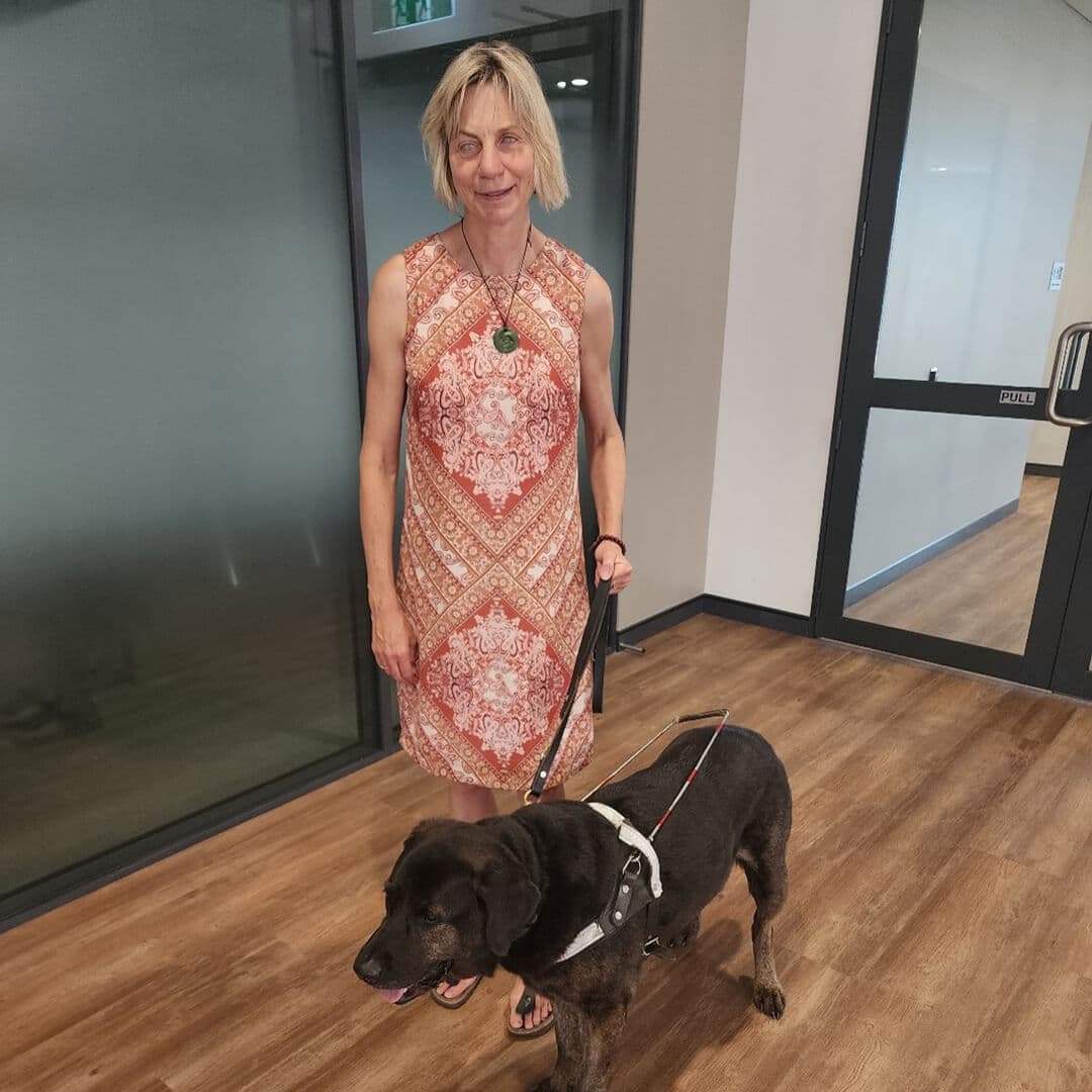 Photo of Maria standing with her guide dog, Mickey. Maria is wearing a patterned dress, and Mickey is in a guide dog harness.