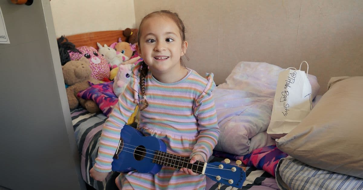 A young child sitting on her bed, smiling, with a ukelele