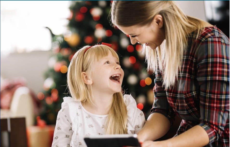 An excited young girl and her mother unwrap a Christmas present with a Christmas tree in the background.