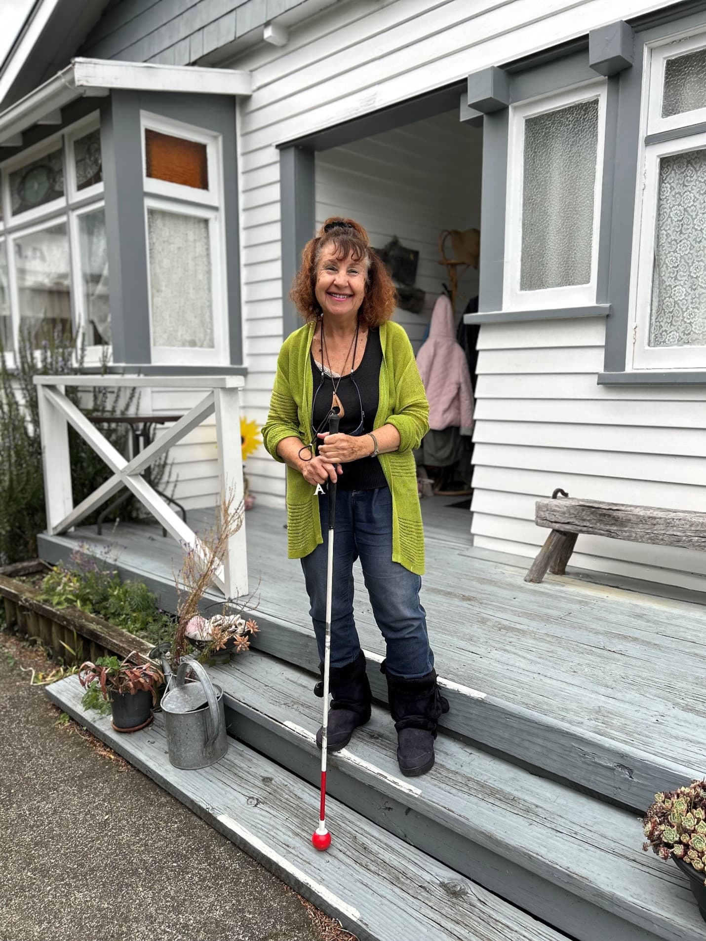 Andrea is standing on some stairs holding her white cane. She is smiling, wearing jeans and a green cardigan.