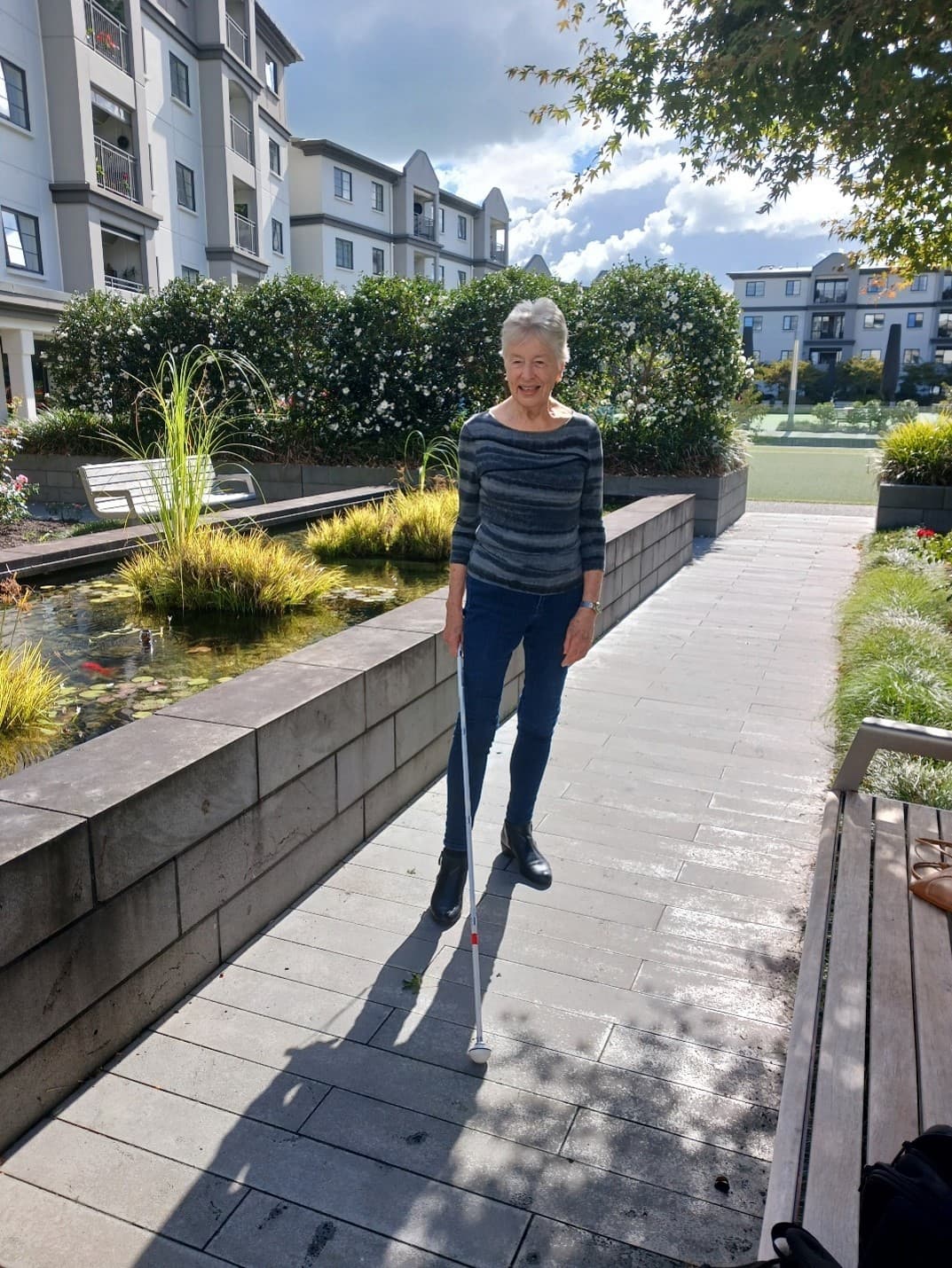 Liz is standing outside in her garden holding her white cane. She is smiling, and wearing black pants with a striped jumper.