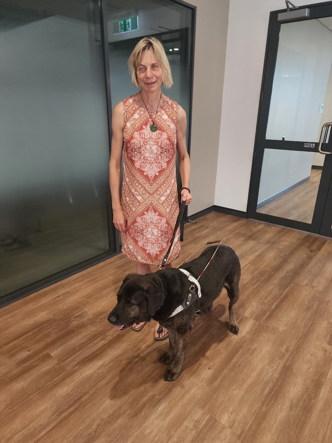 Maria is standing inside with her guide dog Mickey. Maria is wearing a pink and orange patterned dress, and holding Mickey by his harness.
