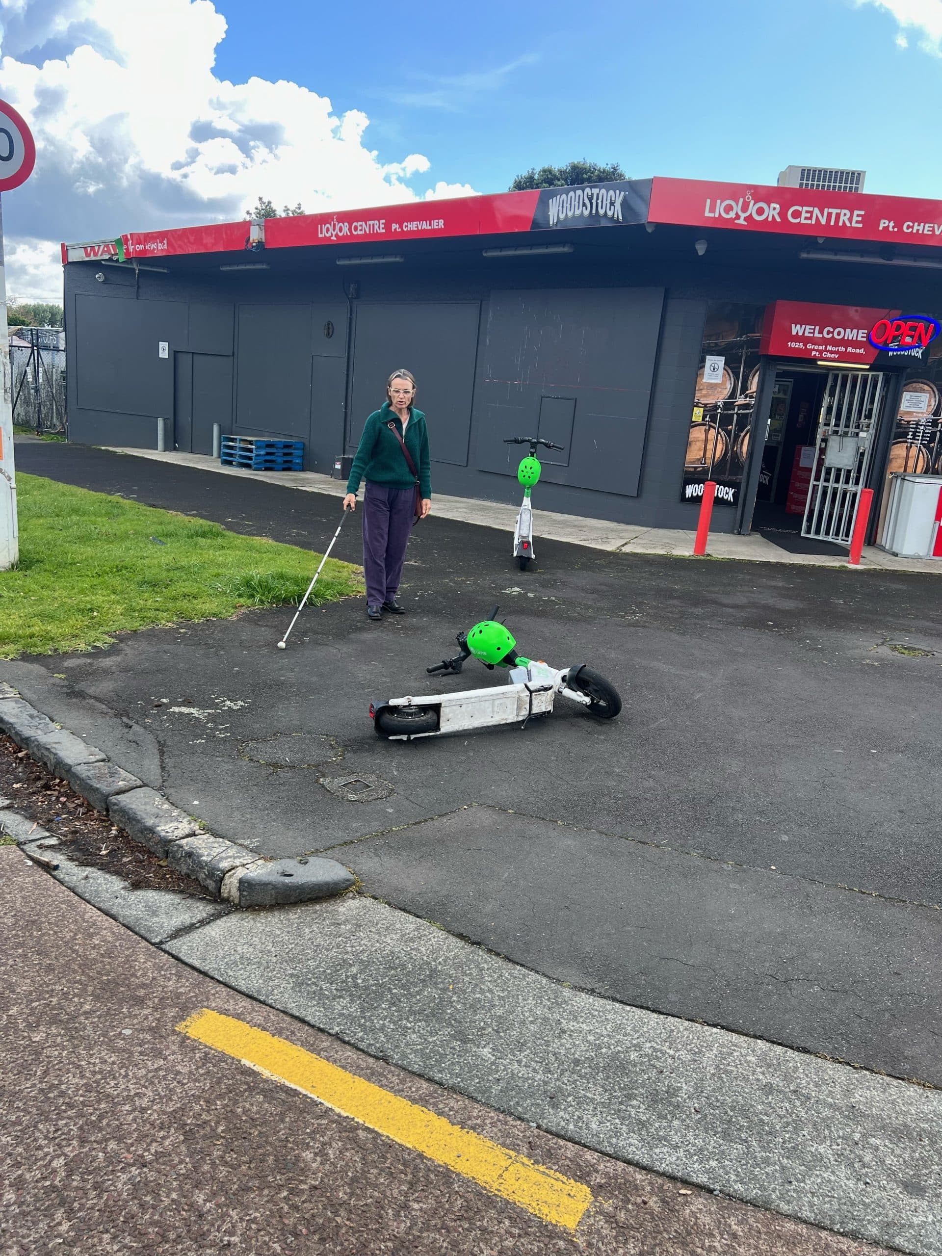 Rhonda is standing outside with her white cane. She is standing behind an e-scooter that has been left on the path.