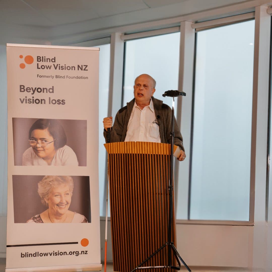 A man standing at a podium, delivering a presentation, behind him is a vertical banner with the Blind Low Vision NZ logo, the text "Beyond Vision Loss", and three pictures of blind and low vision clients.