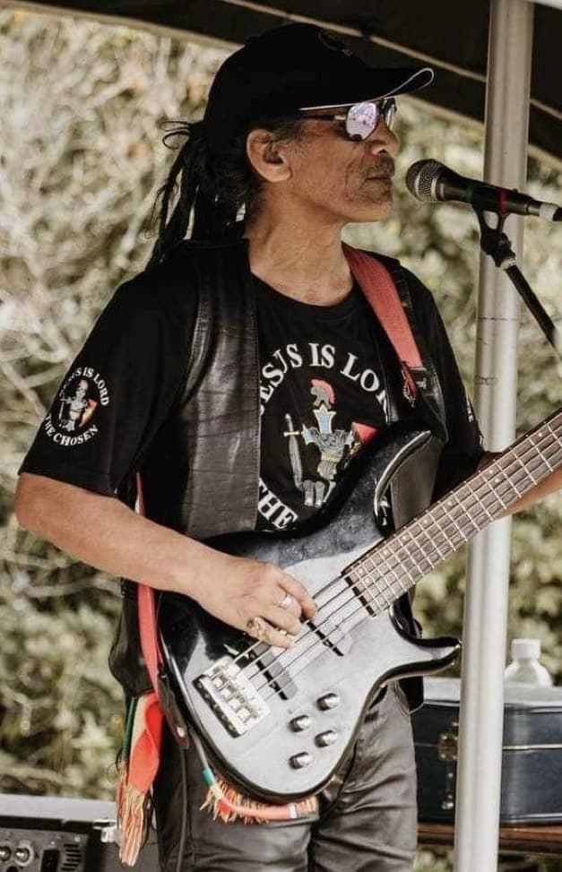 A man with dreadlocks smiling while playing the drums under a tent, holding drumsticks mid-performance.