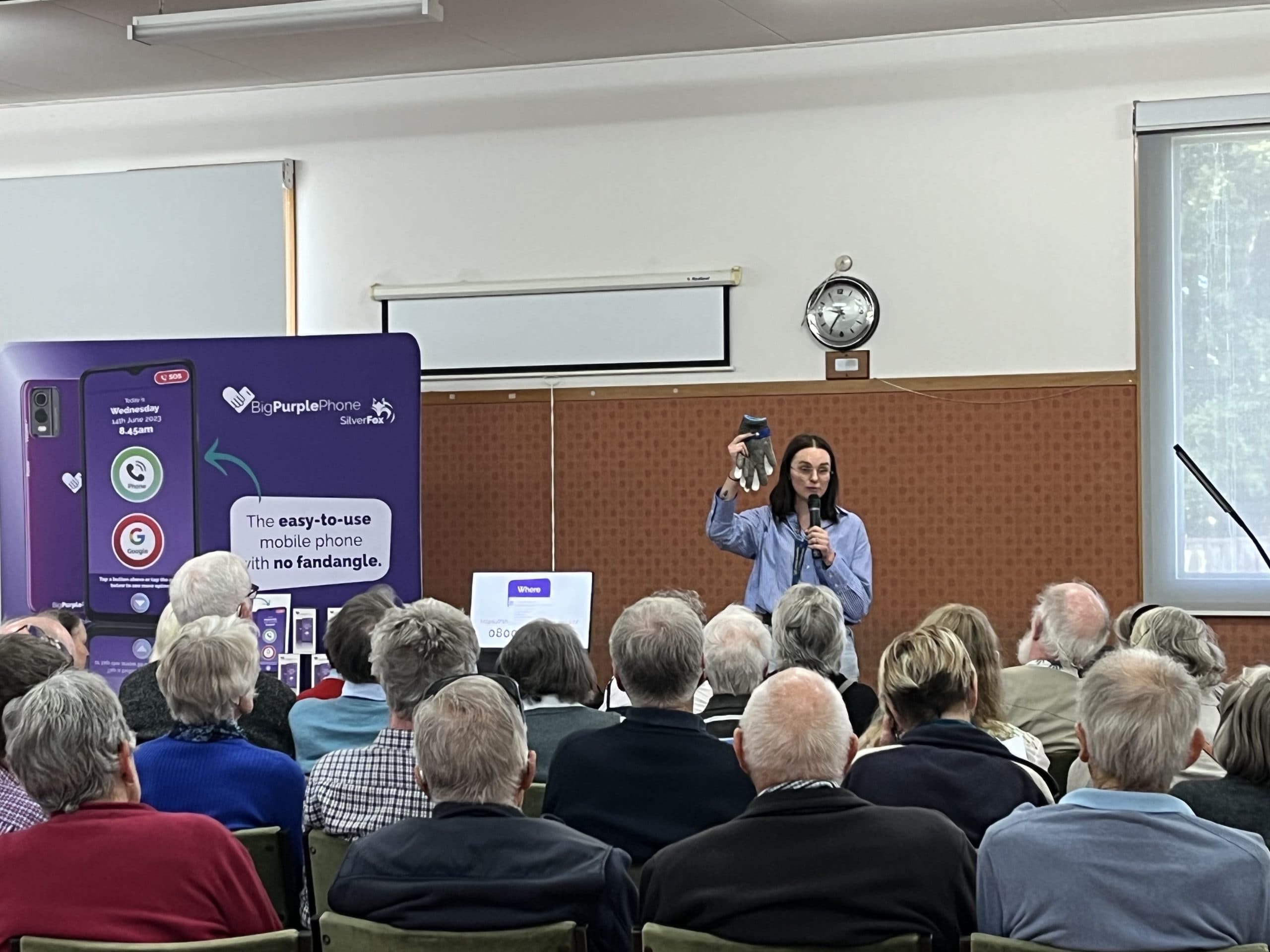 A woman demonstrates a protective glove for people with blindness or low vision to a group of people beside her is an information board promoting the Big Purple Phone from the Vision Store.
