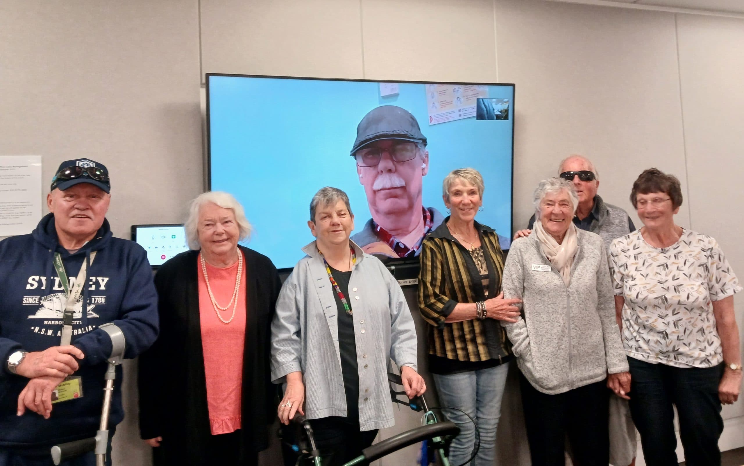 Two men and six women stand in front of a large TV screen featuring a man wearing a flat cap, looking into the camera. They are all smiling at the camera. One man has crutches, while one of the women has a walker.