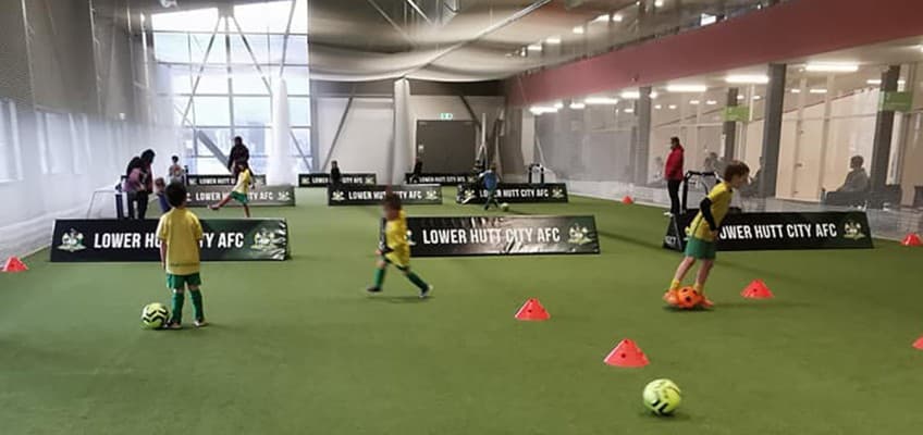 A green indoor space with orange cones, and black banners reading " Lower Hutt City AFC." Three boys wearing bright yellow jerseys and green shorts are practicing dribbling footballs around the orange cones.