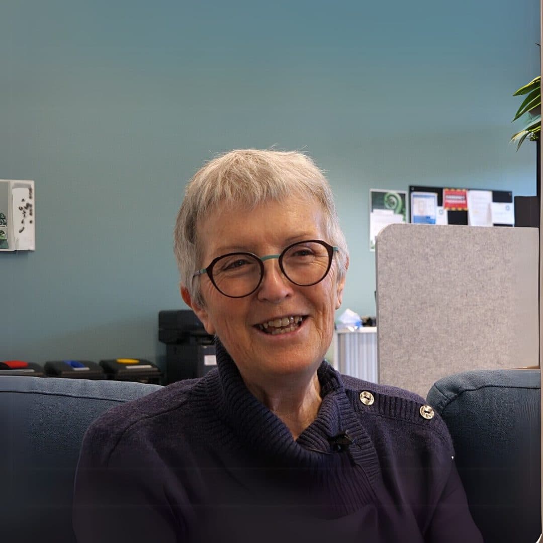 A mature woman with short grey hair in an office wearing a purple jumper with buttons at the shoulder and glasses, smiling for the camera.