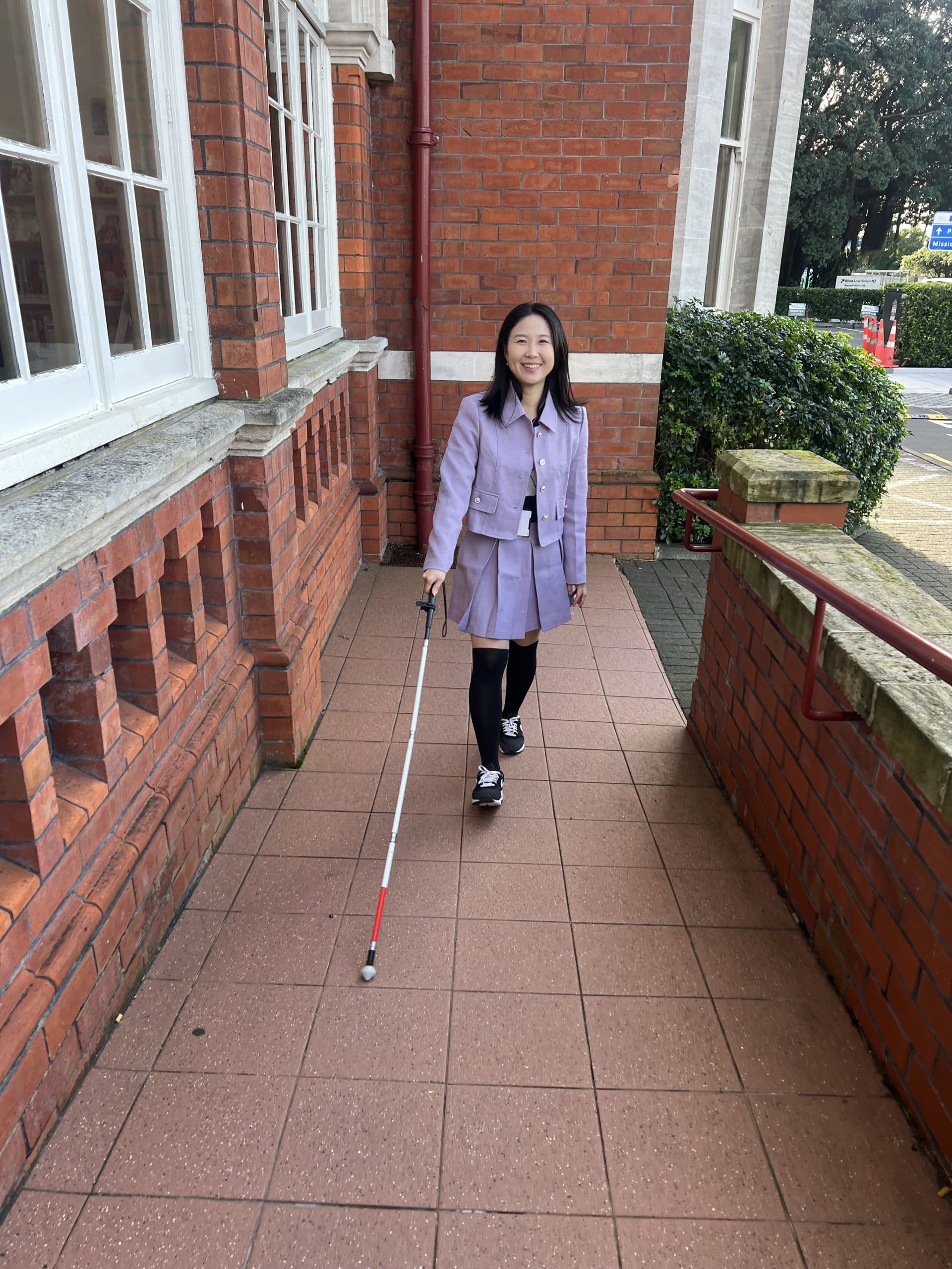 A woman with low vision walks confidently toward the camera outdoors, smiling and using a white cane for navigation.