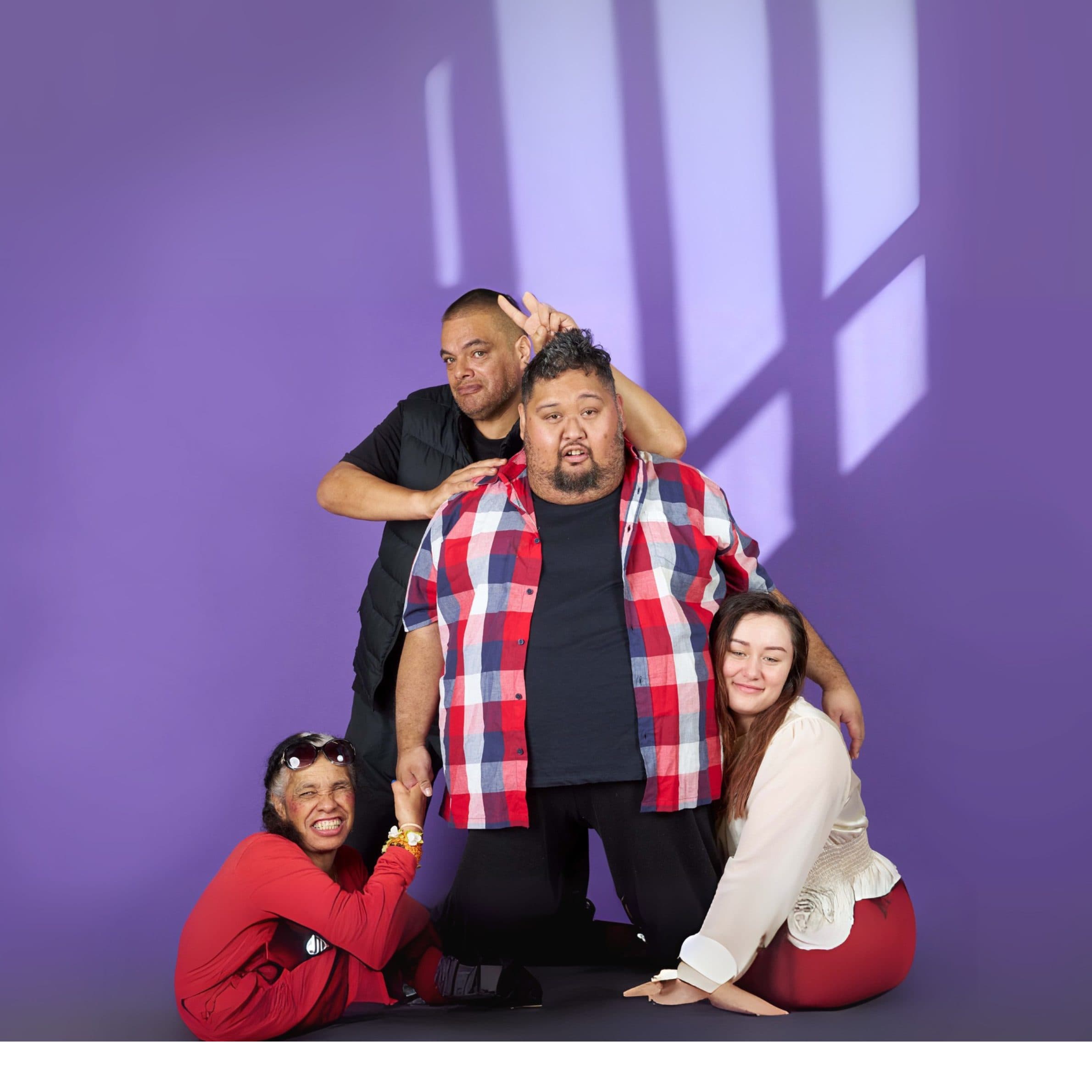 A group of four young people, two men, two women posing for a photo in front of a purple wall. They're grouped closely together with one man at the centre, another man leaning over his shoulder, and two shorter women either side.