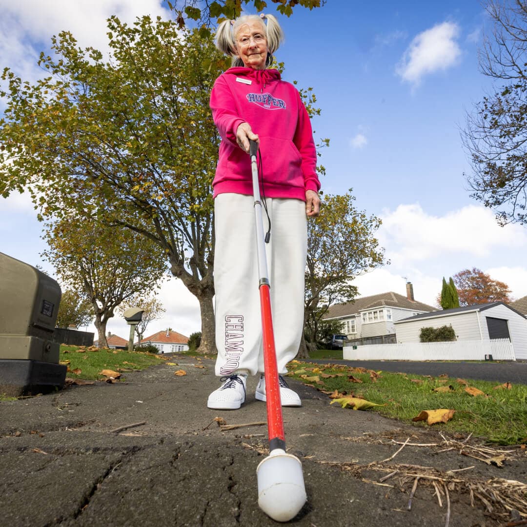 A lady with a white cane standing on a footpath in a suburban setting.