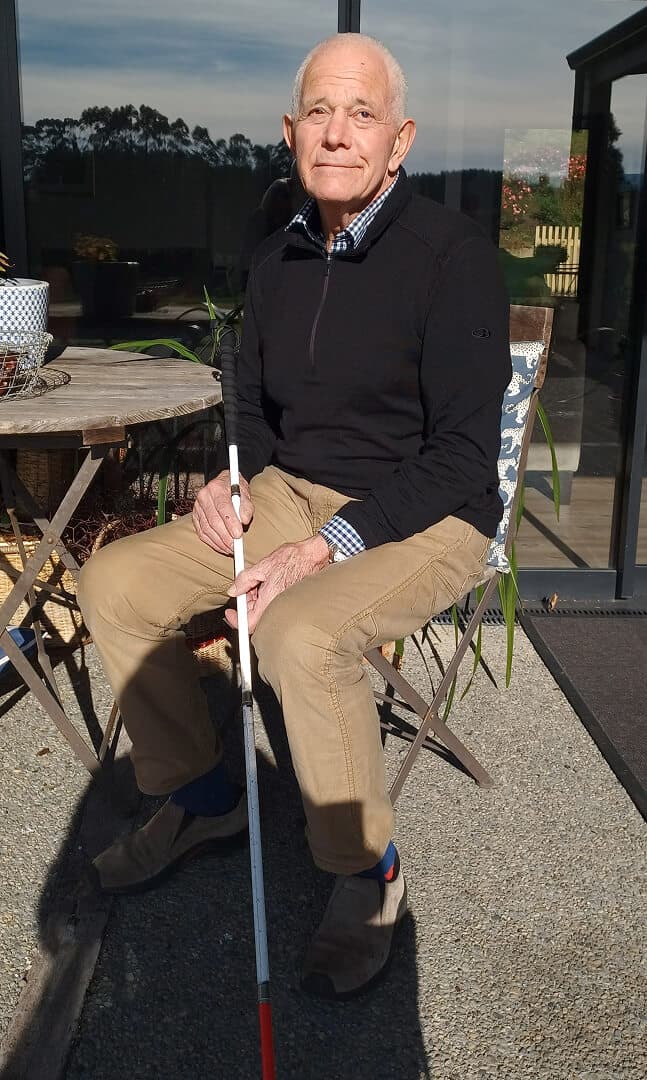 A man sits on a chair outside with his white cane.