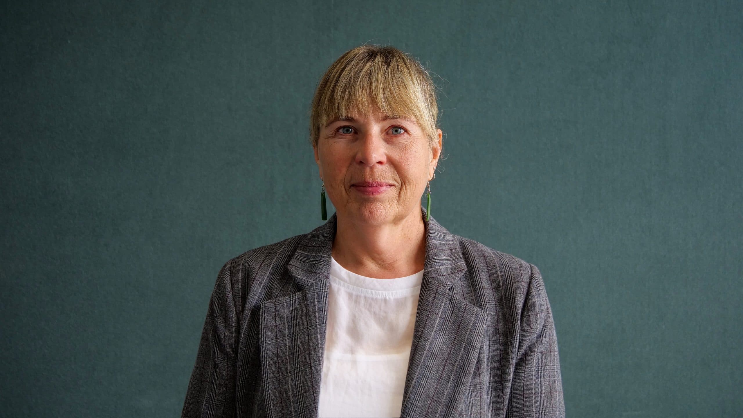 A woman with tied back blonde hair, wearing a grey blazer and a white blouse against a dark grey indoor background smiling for the camera.