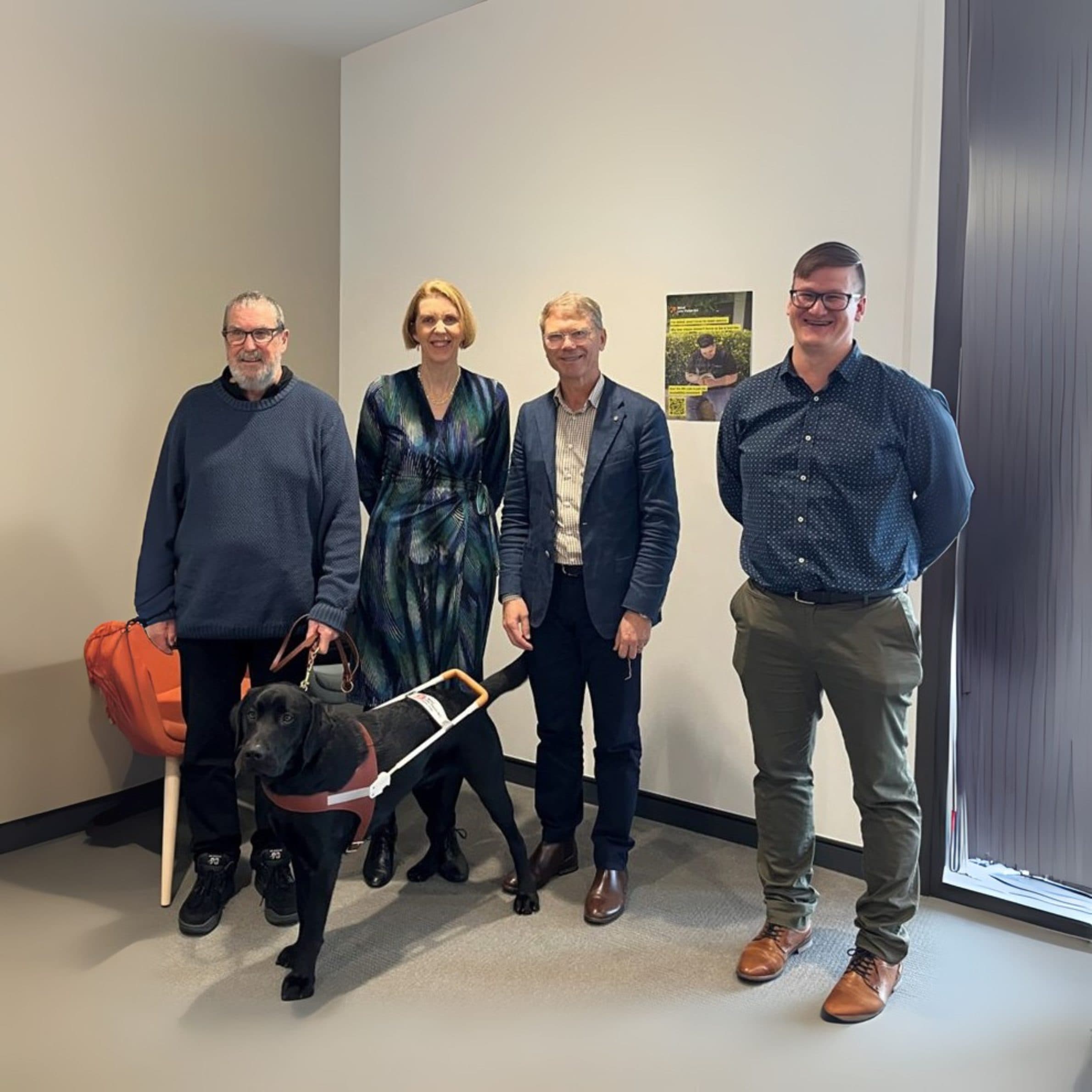 Three men and one woman standing in an office, smiling for the camera with a black lab retriever in a harness.