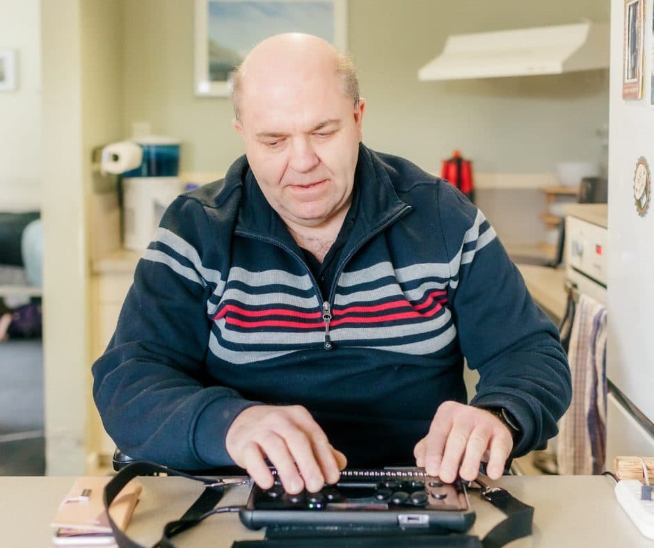 Phil sits at a kitchen counter. He is using a braille device with his hands.