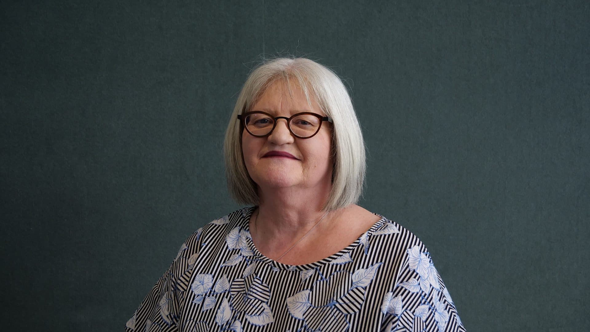 A woman with grey hair wearing glasses, a black, white and blue patterned blouse smiling for the camera with a dark grey internal background.