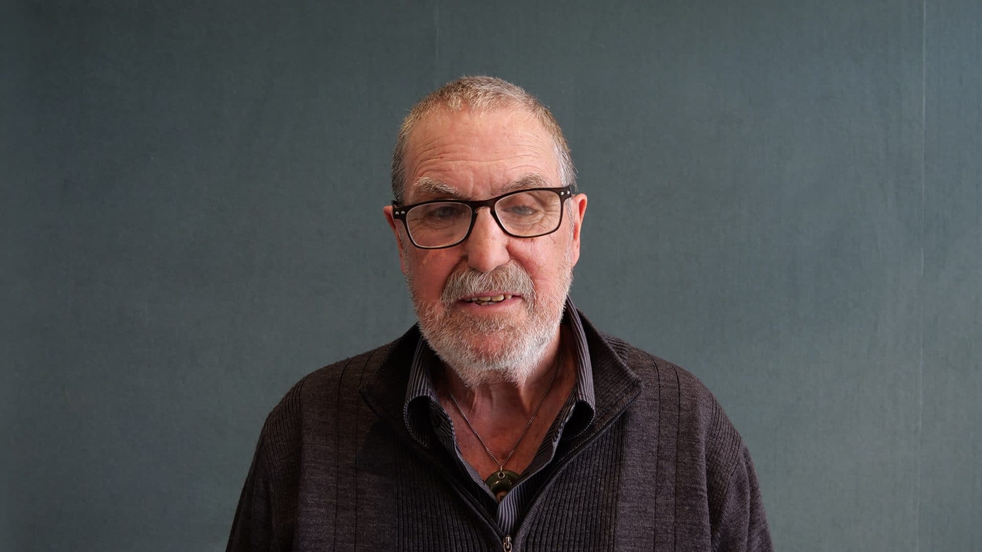 A man with grey hair and a beard wearing glasses, a dark grey zipped jumper and grey striped shirt smiling for the camera with a dark grey internal background.