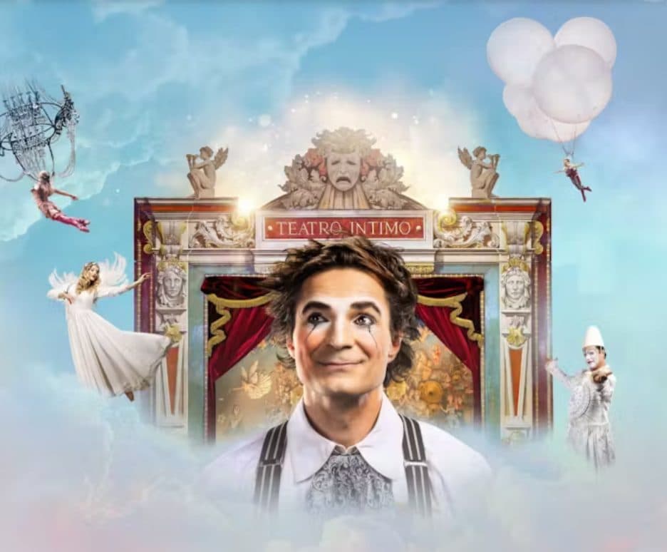 A person in clown makeup, and suspenders smiling with a decorated theatre stage behind him surrounded by an angel, an acrobat suspended from a chandelier, another acrobat hanging from balloons, and a clown.