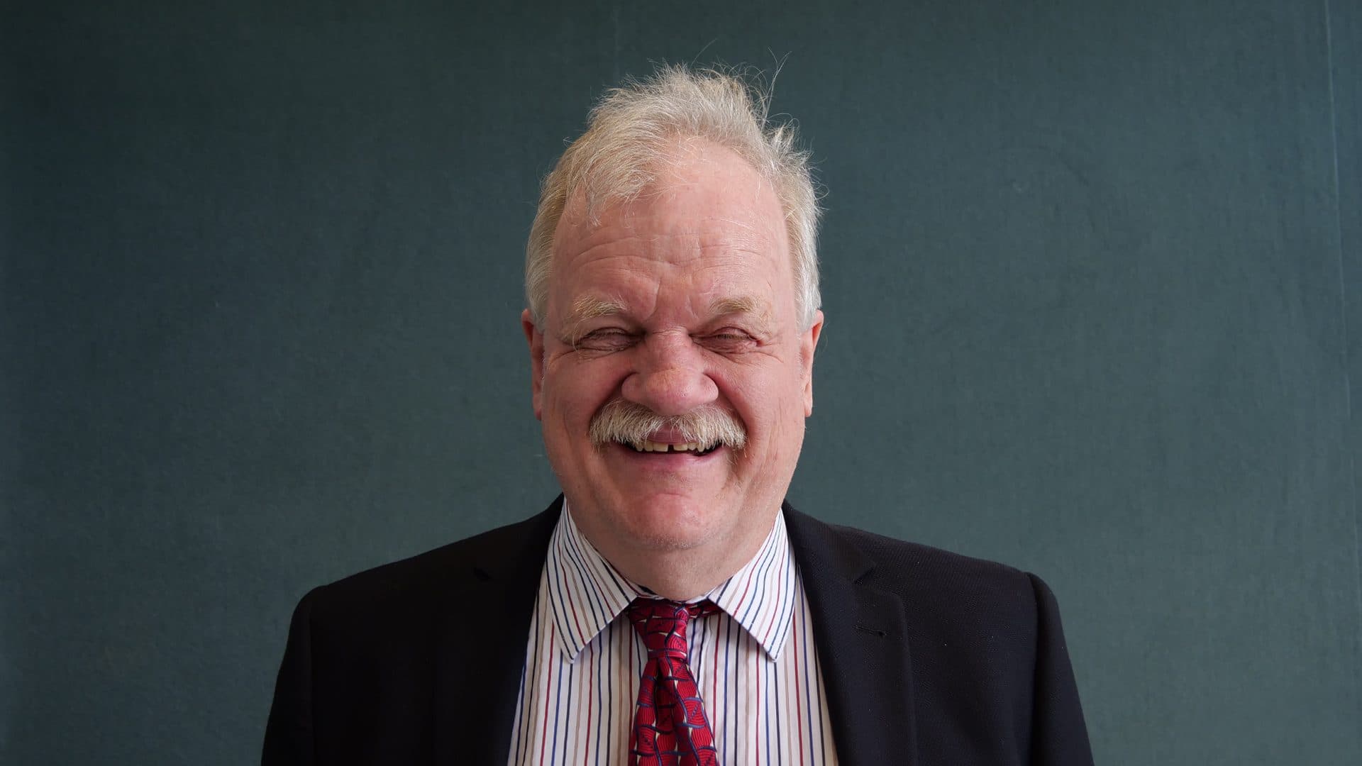 A man with grey hair and a moustache wearing a striped shirt and navy jacket, and red patterned tie smiling for the camera with a dark grey internal background.