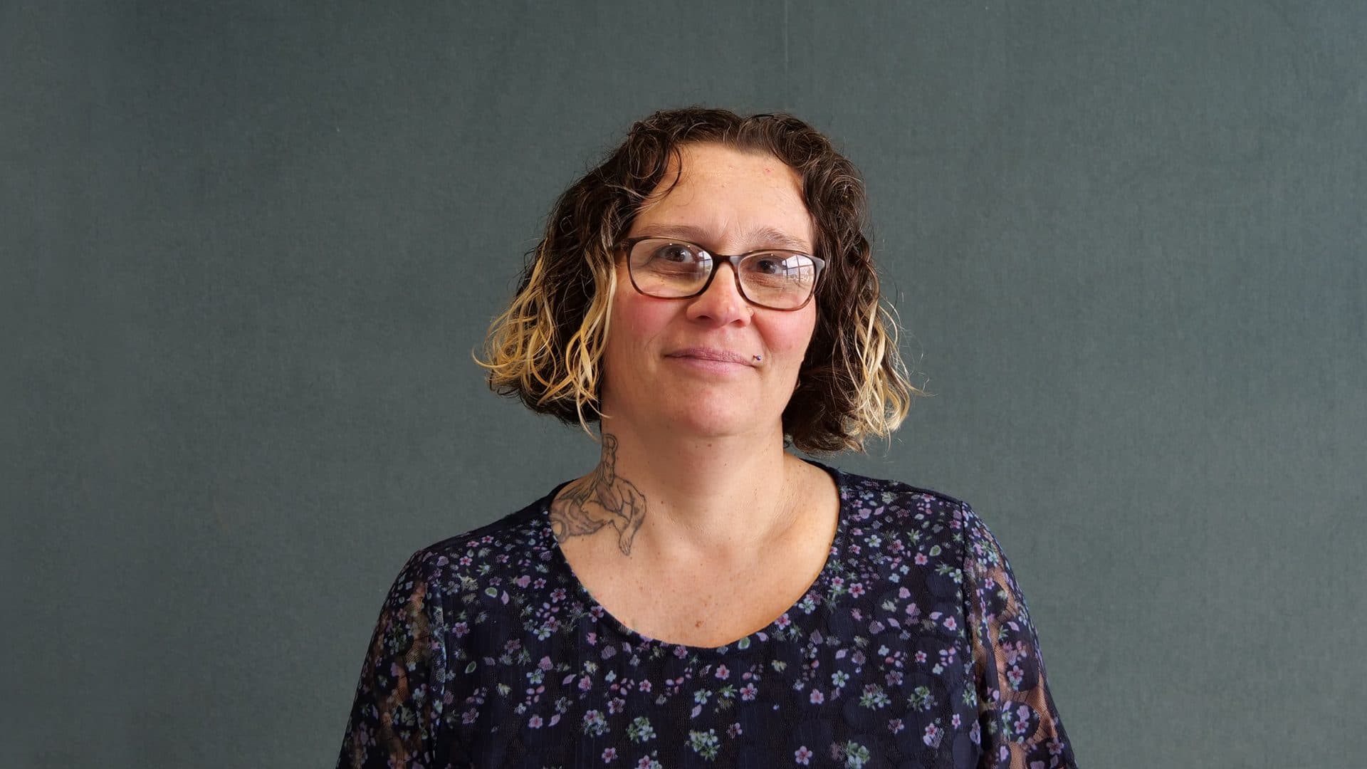 A woman with brown hair wearing glasses, a blue floral patterned shirt smiling for the camera with a dark grey internal background.