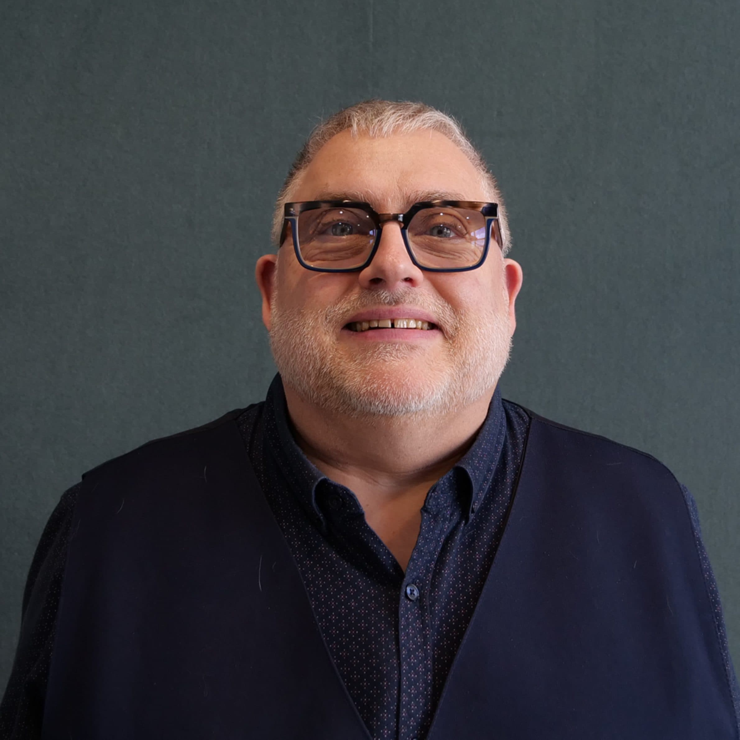 A man with grey hair wearing glasses, a blue patterned shirt and navy vest smiling for the camera with a dark grey internal background.